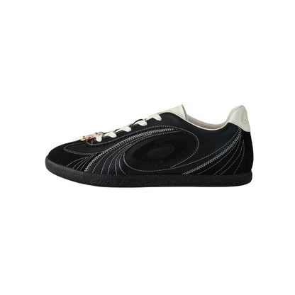 A black low-top sneaker with white laces, white stitching details, and a white heel tab, shown in profile against a solid beige background.