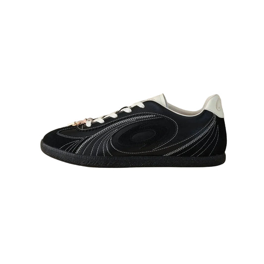 A black low-top sneaker with white laces, white stitching details, and a white heel tab, shown in profile against a solid beige background.