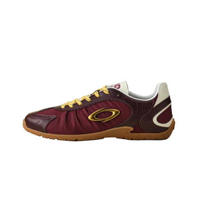 A single Oakley athletic sneaker in burgundy and maroon leather with yellow laces, a gold ring logo, and a brown gum sole, shown in profile against a neutral beige background.