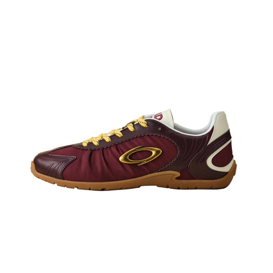 A single Oakley athletic sneaker in burgundy and maroon leather with yellow laces, a gold ring logo, and a brown gum sole, shown in profile against a neutral beige background.