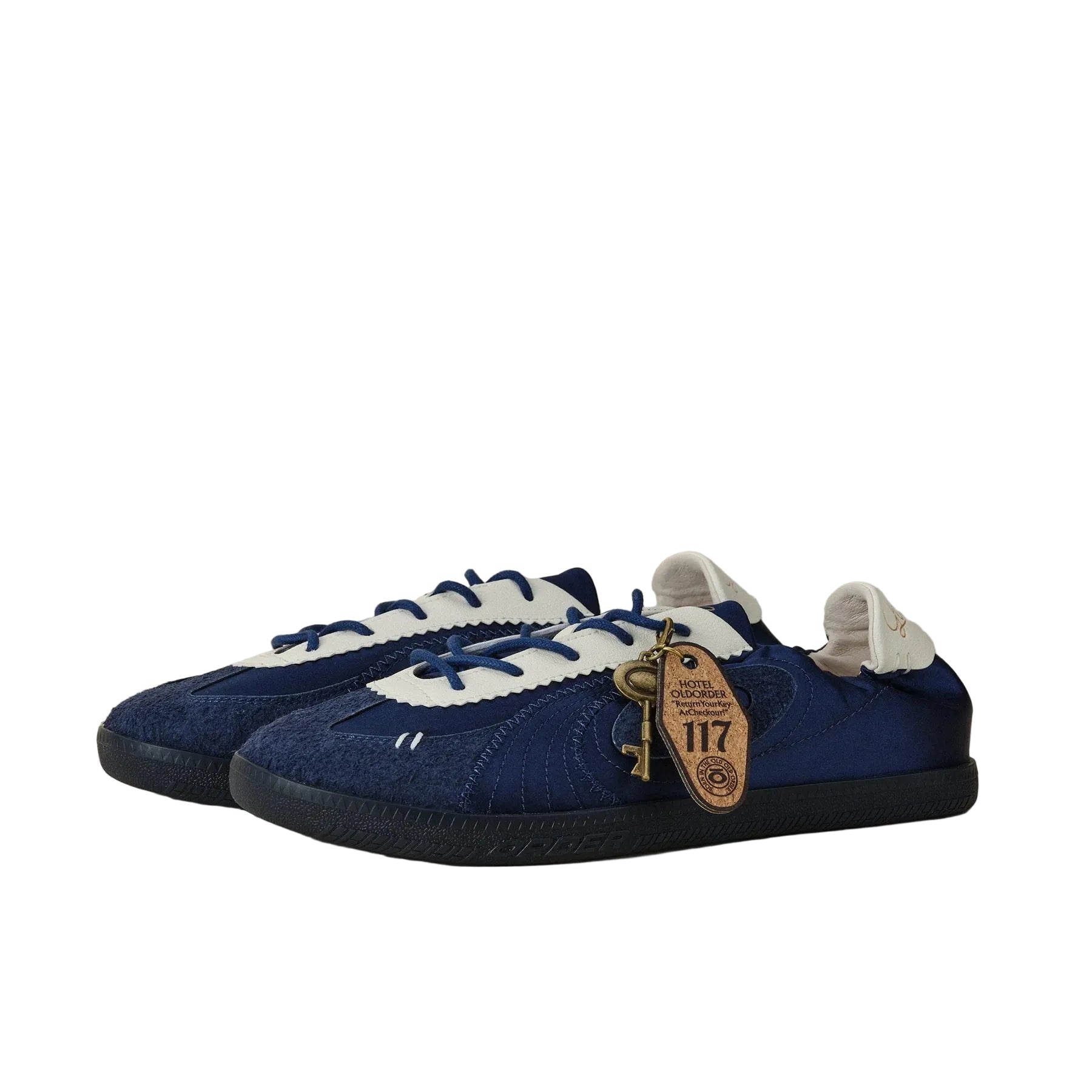 A pair of low-top sneakers in navy blue suede and canvas with white overlays, navy laces, and a brown leather hangtag, photographed on a neutral beige background.